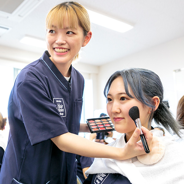 カラフルなパレットを手に持ち、メイクブラシをもう片方の手で学生の頬にあてている女子学生